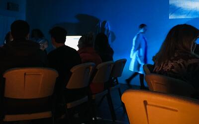 People sitting at tables with screen in a darkened space as someone wearing a lab coat moves between them. Vector used immersive experiences to engage people in the ethical challenges of animal research in news ways.  